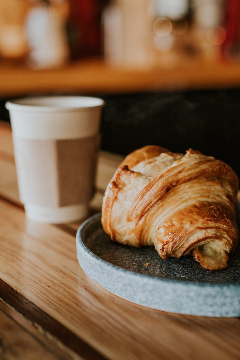 Cozy café setting with paper cup coffee and fresh croissant on a wooden table.